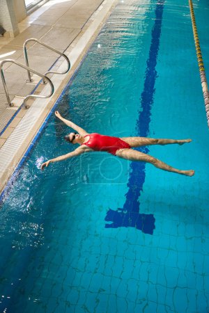 Téléchargez les photos : Maillot de bain féminin sérieux avec bonnet de bain et lunettes flottant à la surface de l'eau avec les bras et les jambes écartées - en image libre de droit