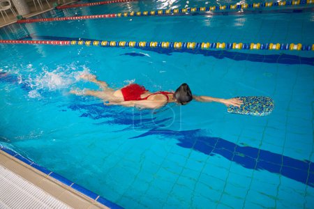 Foto de Deportiva en traje de baño y gorra de natación sosteniéndose en el patinete con la mano mientras flota sobre su estómago en agua - Imagen libre de derechos