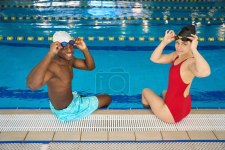 Photo for Joyous interracial swimmers seated on edge of pool putting on goggles before swimming workout - Royalty Free Image