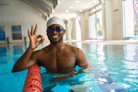 Foto de Retrato de cintura hacia arriba de nadador alegre haciendo signo OK mientras se apoya en divisor de carril en agua turquesa - Imagen libre de derechos