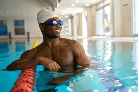 Foto de Sonriente nadador satisfecho en gafas de cian y gorra de natación apoyada en divisor de carril en agua - Imagen libre de derechos