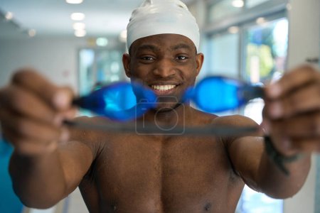 Photo for Portrait of joyful African American swimmer demonstrating pair of swim goggles before workout - Royalty Free Image