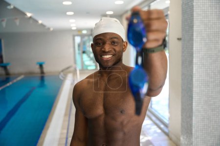 Photo for Waist-up portrait of joyful sportsman showing off pair of swimming goggles while standing poolside - Royalty Free Image