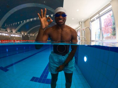 Foto de Alegre nadador masculino en gorra de natación y gafas haciendo signo OK mientras está de pie en la piscina - Imagen libre de derechos