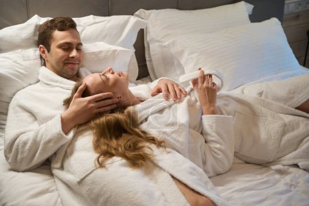 Téléchargez les photos : Couple heureux amoureux dans des peignoirs couchés dans un lit confortable et parlant, rêvant de grandes vacances, appréciant l'intérieur et le service de la chambre d'hôtel - en image libre de droit