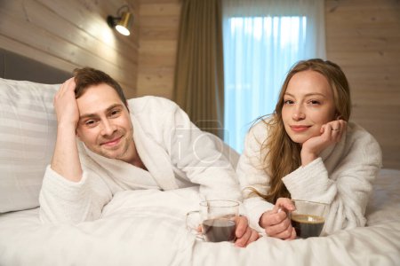 Téléchargez les photos : Beau couple tout juste marié profitant d'une lune de miel couché sur le lit dans des peignoirs blancs moelleux et boire du café après les procédures de spa, passe-temps récréatif - en image libre de droit