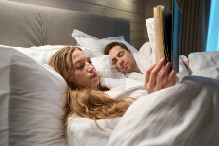 Foto de Esposo durmiendo acostado cerca de su esposa que yace en albornoz y leyendo un libro interesante hasta tarde, pareja descansando después de un día de trabajo agotado - Imagen libre de derechos