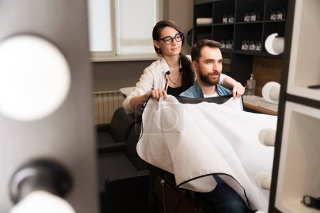 Téléchargez les photos : Barber femme enlève la cape de protection du client, les gens sont reflétés dans le miroir - en image libre de droit