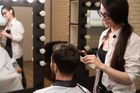 Téléchargez les photos : Jeune femme barbier clipping mâle, maître et client sont reflétés dans le miroir - en image libre de droit