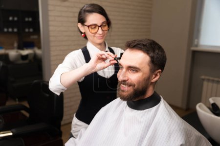 Foto de Artesana en gafas y un pendiente rojo corta el cabello de un cliente con tijeras, un hombre con una capa protectora - Imagen libre de derechos