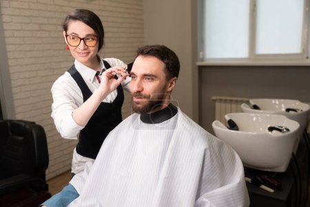 Téléchargez les photos : Jeune coiffeur coupe un client cheveux avec des ciseaux, à côté d'un évier spécial pour laver les cheveux - en image libre de droit