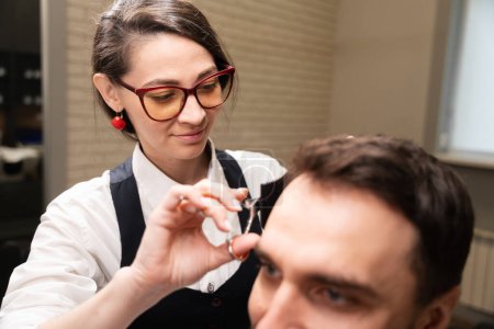 Téléchargez les photos : Jeune femme avec une boucle d'oreille rouge coupe un client cheveux avec des ciseaux, - en image libre de droit