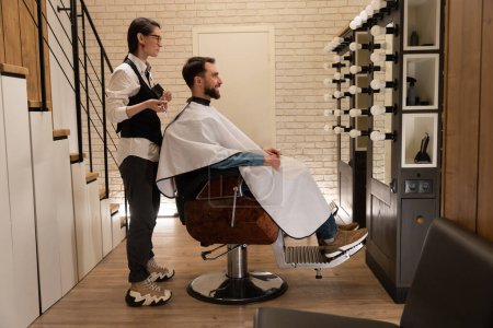 Foto de Cliente satisfecho se sienta en una barbería en una silla de peluquería, al lado de un maestro con tijeras - Imagen libre de derechos