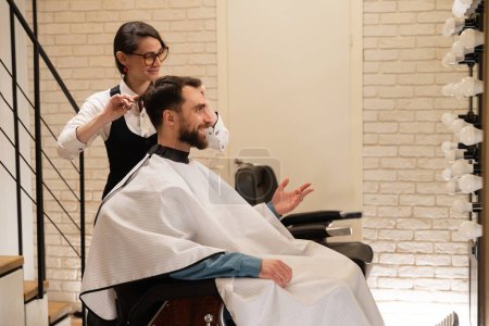 Foto de Cliente sonriente se sienta en una barbería en una silla de peluquería, una mujer le hace un corte de pelo con tijeras - Imagen libre de derechos