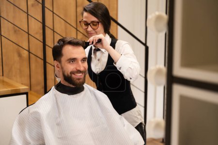 Téléchargez les photos : Un client souriant s'assoit dans une chaise de coiffeur en cape de protection, le coiffeur le coupe avec des ciseaux - en image libre de droit