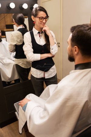 Foto de La maestra se comunica con un cliente barbudo, un hombre se sienta frente a un espejo - Imagen libre de derechos