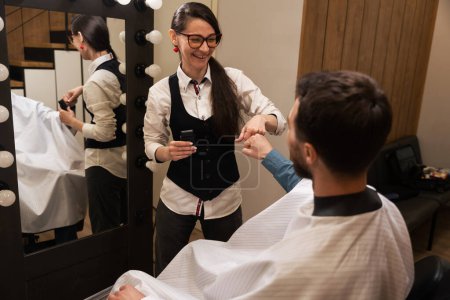 Foto de Artesana alegre en la barbería da la bienvenida al cliente, el hombre se sienta frente al espejo - Imagen libre de derechos