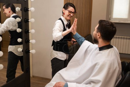 Foto de Mujer en un peluquero en una peluquería da la bienvenida a un cliente, un hombre se sienta en una silla de barbero - Imagen libre de derechos