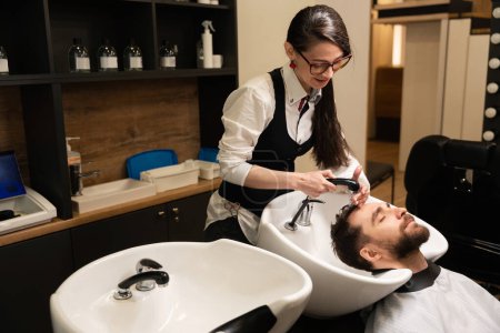 Téléchargez les photos : Jeune femme coiffeur lave les cheveux d'un homme, le client est situé à un évier spécial - en image libre de droit