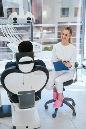 Téléchargez les photos : Femme confiante stomatologue tenue de dossiers médicaux à l'écoute du patient et assis dans un bureau de luxe avec un équipement moderne, service de clinique privée - en image libre de droit