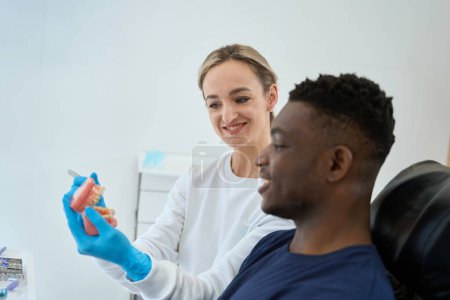 Téléchargez les photos : Femme souriante technicienne dentaire montrant une prothèse dentaire à un client afro-américain, un médecin et un patient discutant de variantes de prothèses dentaires - en image libre de droit