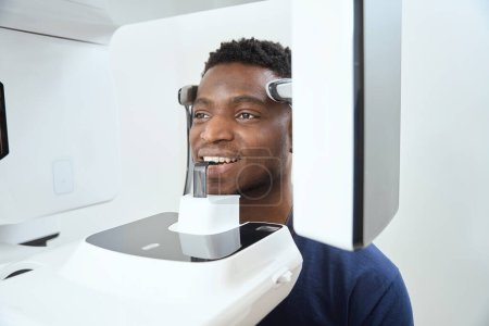 Téléchargez les photos : Homme américain assis à la clinique près de l'appareil de tomographie informatique. Image tomographique du crâne et des dents - en image libre de droit