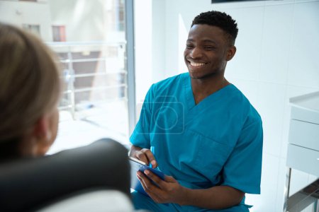 Téléchargez les photos : Homme souriant en costume bleu tenant la tablette et parlant à la femme, regardant la femme dans la stomatologie - en image libre de droit