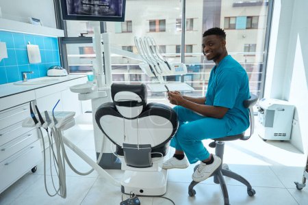 Téléchargez les photos : Jeune médecin en peignoir bleu assis sur une chaise près de l'équipement et tenant le téléphone, regardant la caméra - en image libre de droit