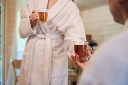 Photo for Close up of woman in dressing gown holding and giving cups of tea to man - Royalty Free Image