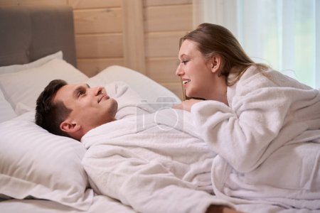 Téléchargez les photos : Femme souriante allongée sur un homme heureux portant des robes de chambre blanches tout en restant au lit - en image libre de droit