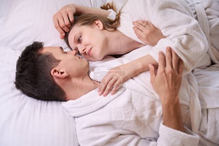 Téléchargez les photos : Homme et femme souriants en robes de chambre se regardant tout en étant couchés au lit - en image libre de droit