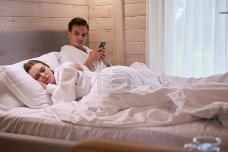 Téléchargez les photos : Homme tenant smartphone dans les mains et regardant dame endormie tout en étant couché au lit - en image libre de droit