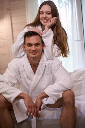 Foto de Sonriente joven hombre y mujer en batas blancas sentados en el borde de la cama - Imagen libre de derechos
