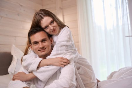 Téléchargez les photos : Femme heureuse en peignoir étreignant l'homme heureux par le cou et se blottissant par derrière - en image libre de droit