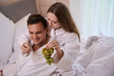 Téléchargez les photos : Femme portant une robe de chambre et offrant un homme souriant avec des raisins au lit - en image libre de droit