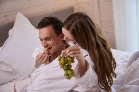 Foto de Mujer vistiendo bata y alimentando al hombre feliz con uvas en la cama - Imagen libre de derechos