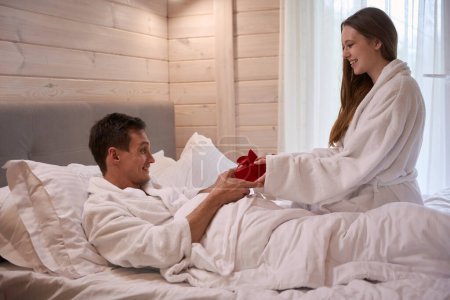 Téléchargez les photos : Dame heureuse portant un peignoir et donnant un cadeau à un homme souriant couché dans son lit - en image libre de droit