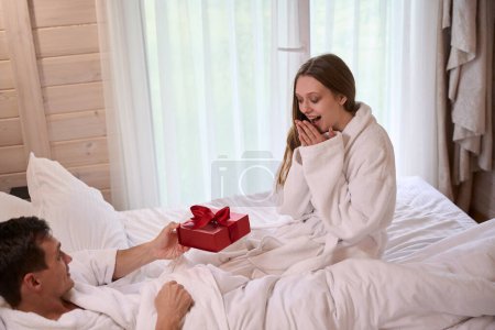 Foto de Mujer feliz usando albornoz y dando regalo a hombre sonriente acostado en la cama - Imagen libre de derechos