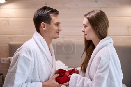 Téléchargez les photos : Heureux homme et femme en peignoirs se regardant tout en tenant boîte-cadeau rouge - en image libre de droit