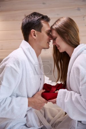 Téléchargez les photos : Heureux homme et femme en peignoirs penchant leur front tout en tenant boîte cadeau rouge - en image libre de droit