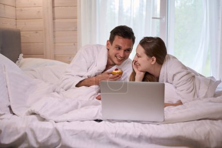 Téléchargez les photos : Couple en peignoirs couché dans le lit avec ordinateur portable tandis que l'homme tenant gâteau - en image libre de droit