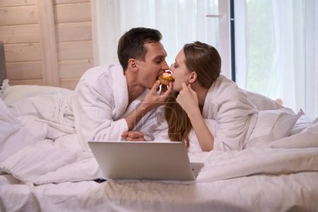 Foto de Pareja en albornoces acostados en la cama con el ordenador portátil mientras comen pastel juntos - Imagen libre de derechos