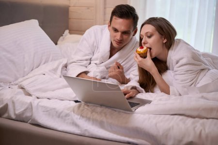 Téléchargez les photos : Couple en peignoirs couché au lit avec ordinateur portable pendant que la femme mange du gâteau - en image libre de droit