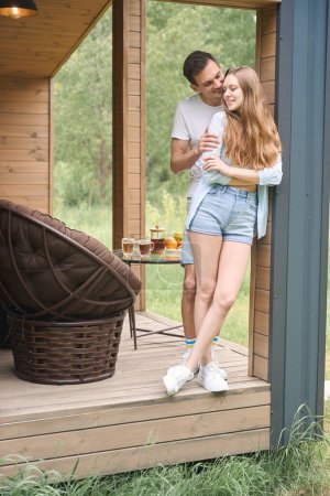 Téléchargez les photos : Homme heureux embrassant dame par derrière tout en se tenant sur la terrasse de la maison de campagne - en image libre de droit