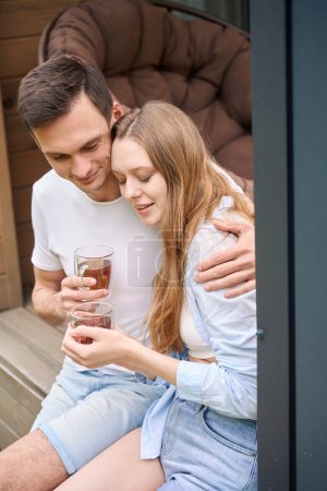 Téléchargez les photos : Homme étreignant la femme par les épaules tout en étant assis sur la terrasse du chalet et tenant des tasses à thé - en image libre de droit