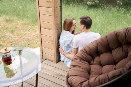 Foto de Sonriente pareja amorosa hablando y mirándose mientras están sentados en el borde de la terraza - Imagen libre de derechos