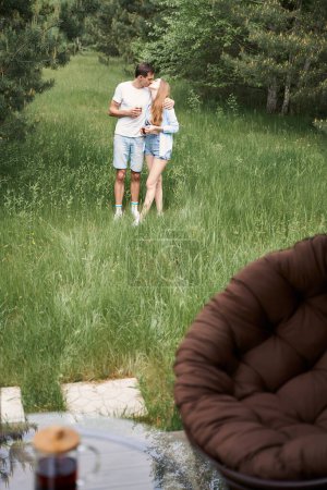 Téléchargez les photos : Homme et femme heureux tenant des tasses à thé et embrasser tout en se tenant dans la prairie près de la maison de campagne - en image libre de droit