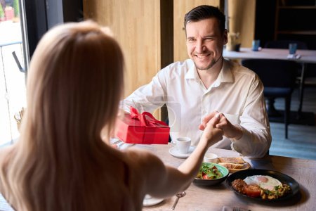 Téléchargez les photos : Beau souriant homme faisant cadeau à sa femme pendant le petit déjeuner dans un café confortable, femme acceptant avec gratitude le cadeau de son bien-aimé, vue de dos - en image libre de droit