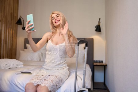Photo for Happy blond woman blogger making selfie or streaming video sitting on bed of hotel suite, talking to her followers, making room tour arriving to vacation - Royalty Free Image