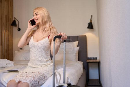 Foto de Mujer rubia alegre hablando teléfono sentado en la cama y sosteniendo la maleta, llamando al taxi para llegar al aeropuerto, dejando el hotel, fin de vacaciones - Imagen libre de derechos